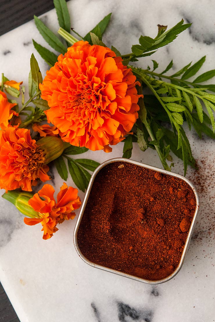 A container of chipotle spice next to a few marigold flowers on a marble tray.
