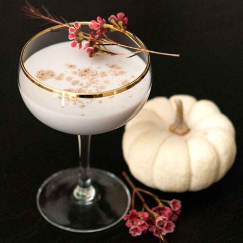 A White Pumpkin cocktail on a table, garnished with a few flowers beside a small white pumpkin on a table.