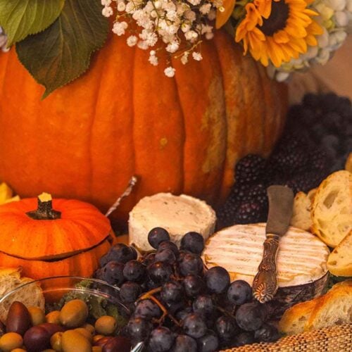 A pumpkin filled with flowers sitting in a tray full of cheese, grapes, bread, and olives with a small pumpkin gourd.
