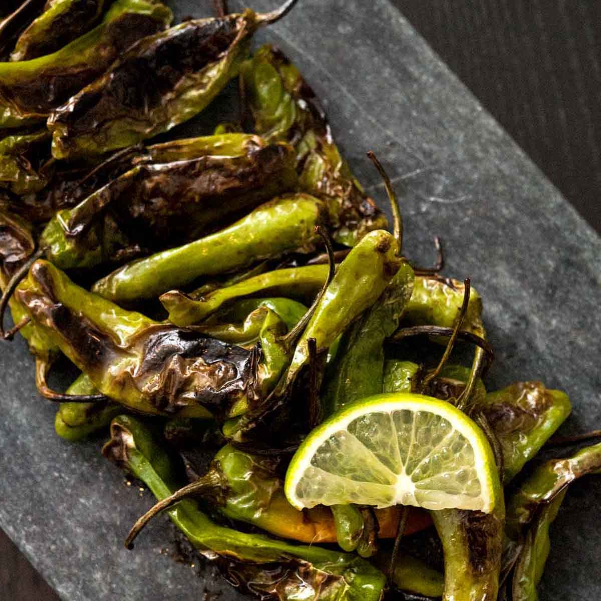 Blistered shishito peppers on a slate with a slice on lime.