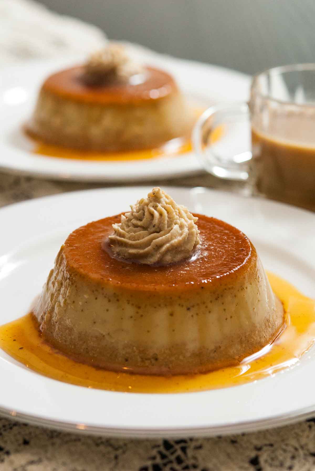 2 pumpkin flans on plates on a table, topped with chai whipped cream, with a small cup of coffee.