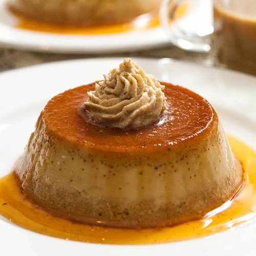 2 pumpkin flans on plates on a table, topped with chai whipped cream, with a small cup of coffee.