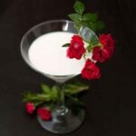 A White Rosemary Rose cocktail in a martini glass, garnished with small roses, with a few roses at its base.