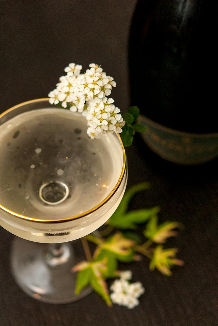 A cocktail garnished with flowers on a black table, sprinkled with small green leaves. A prosecco bottle in the background.