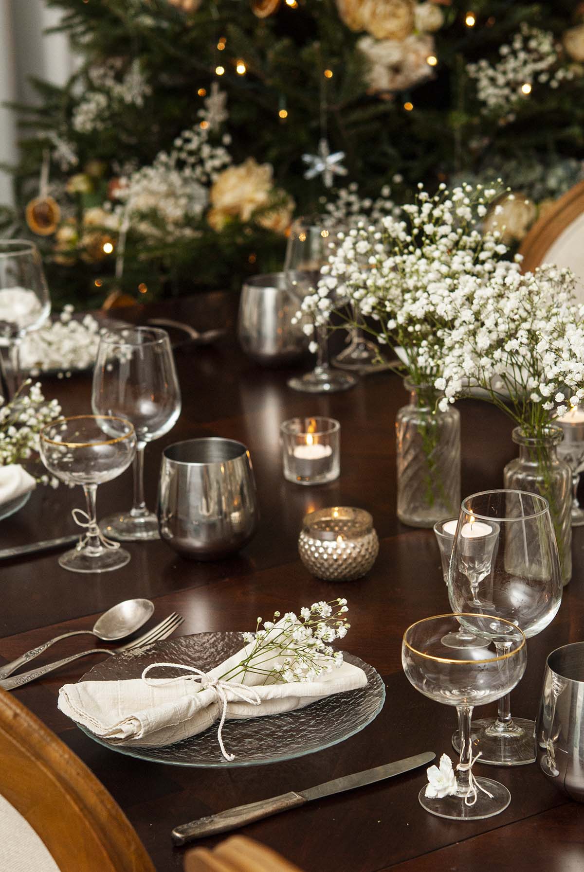 A decorated dinner table with baby's breath in front of a decorated Christmas tree.