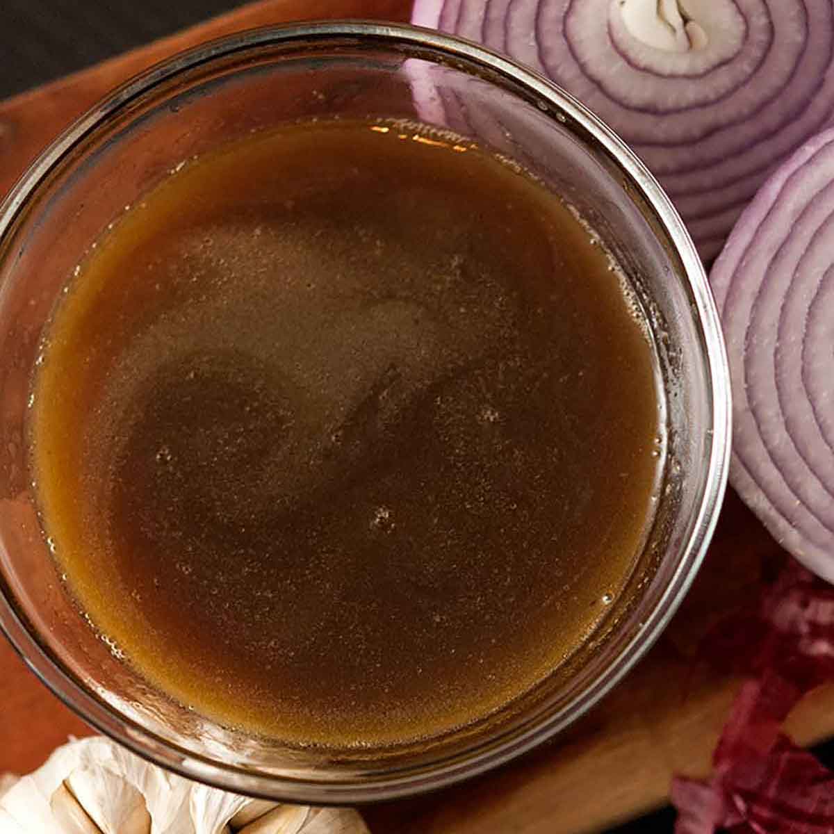 A bowl of bone broth on a wooden board with onions and garlic.