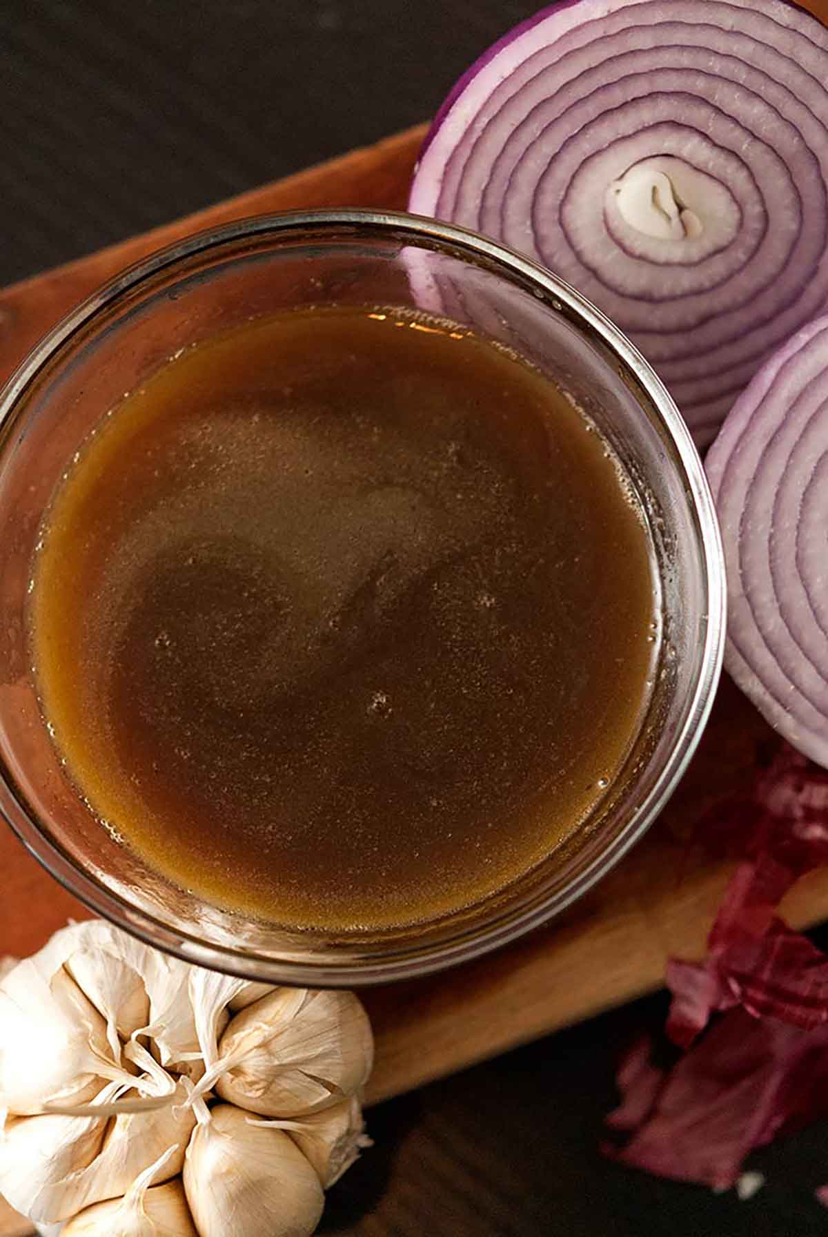 This beef bone broth is a fantastic base for rich soup recipes or simply sipping for better health. #bonebroth, #recipe A bowl of bone broth on a wooden board with onions and garlic.
