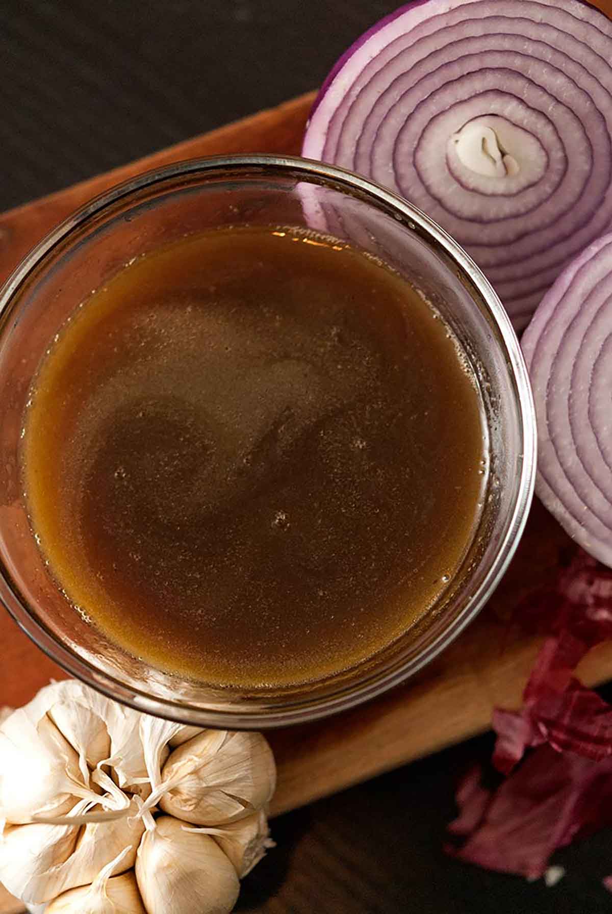 A bowl of bone broth on a wooden board with onions and garlic.