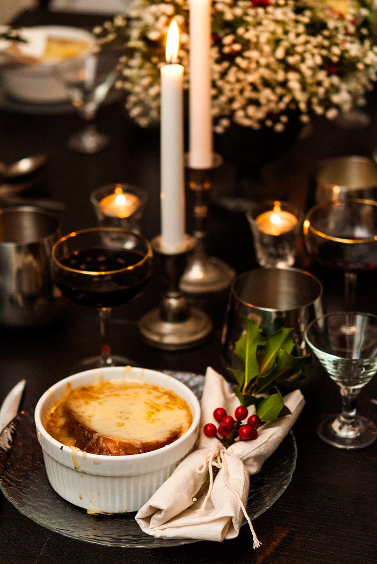 French onion soup on a table lit with candles, surrounded by glassware and Christmas holly decorations.