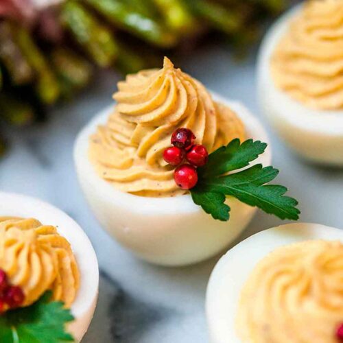 A deviled egg garnished with pink peppercorns and parsley, surrounded by other deviled eggs and asparagus.