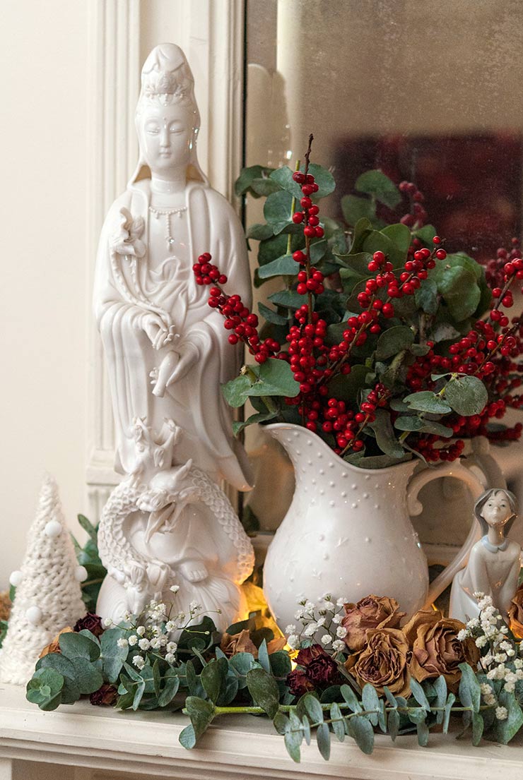 A Buddha statue on a mantle surrounded by flowers, tree decoration and a white jug of berries with leaves.