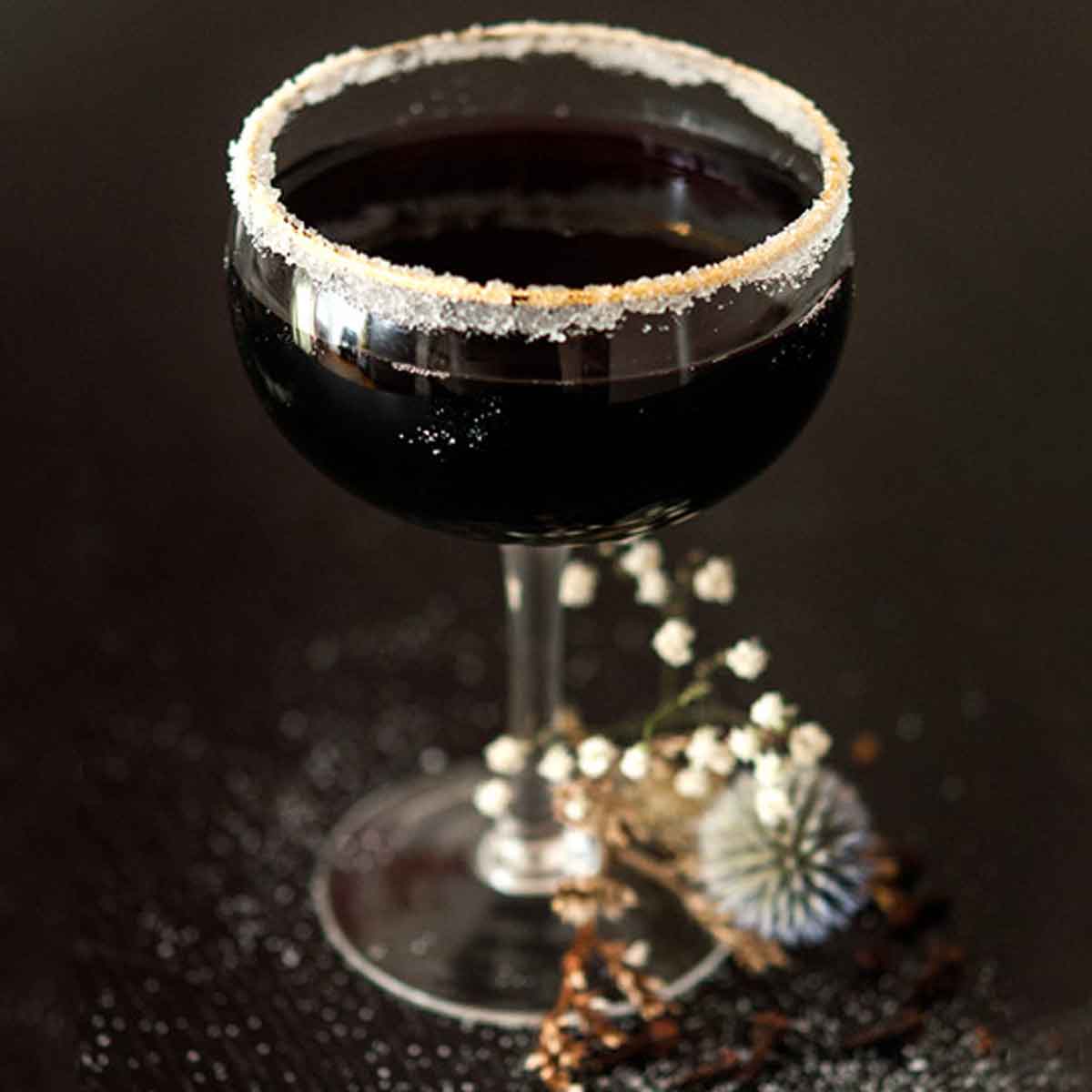 A sugar-rimmed, black cocktail surrounded by dry baby's breath on a sugar-sprinkled table.
