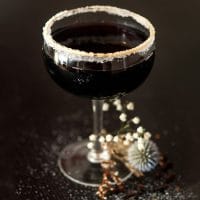 A haunting black cocktail in a coup glass, rimmed with sugar, with dry flowers at its base.