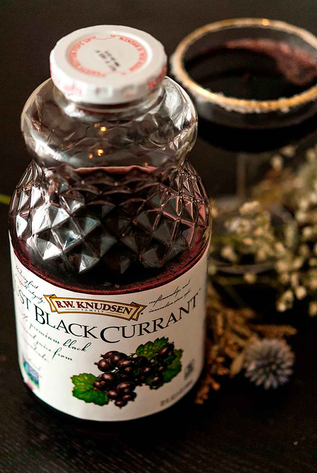 A bottle of black currant juice in front of a sugar-rimmed cocktail and dry baby's breath flowers.