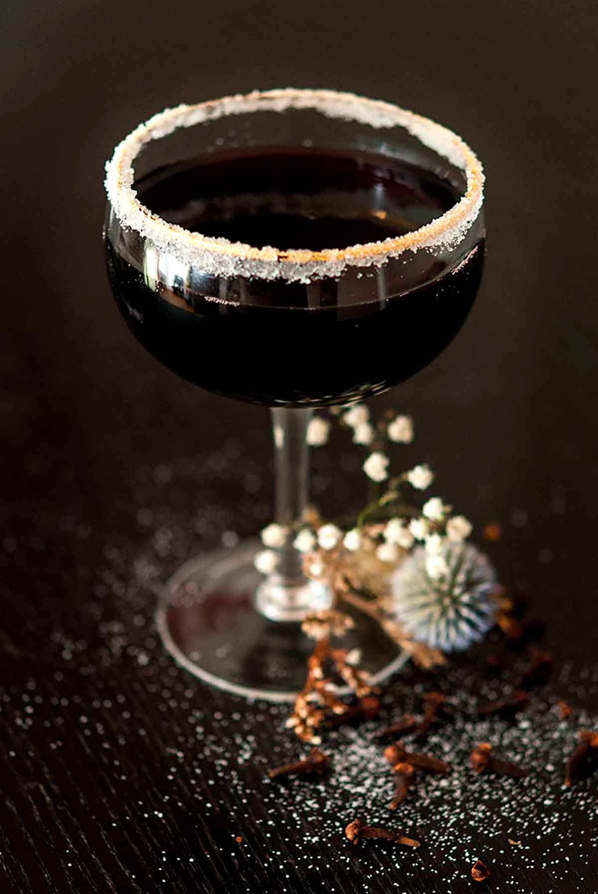 A sugar-rimmed, black cocktail surrounded by dry baby's breath on a sugar-sprinkled table.