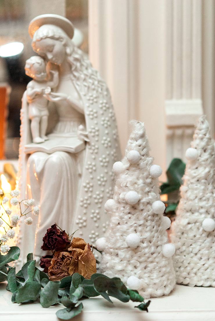 Small Christmas trees made of yarn on a mantle decorated with dry roses, eucalyptus and a porcelain Mary statue.