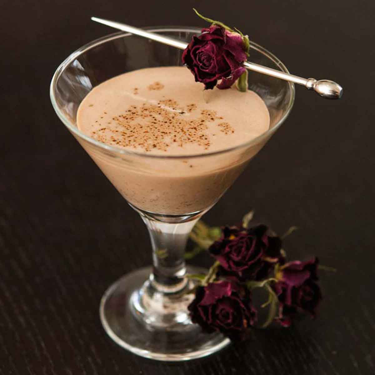 A small glass of Mexican Chocolate liqueur on a table, garnished with a dry rose with 3 roses at its base.