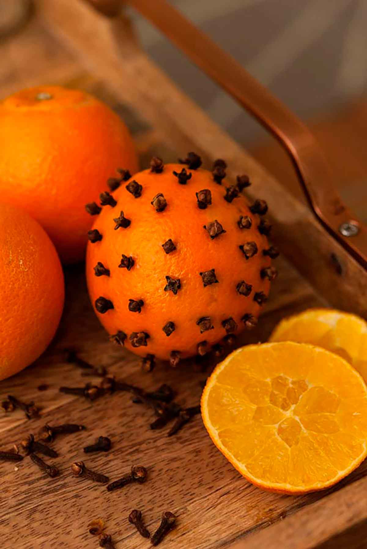 An orange with cloves stuck evenly into it on a wooden tray, surrounded by a 2 more oranges and 2 orange slices.