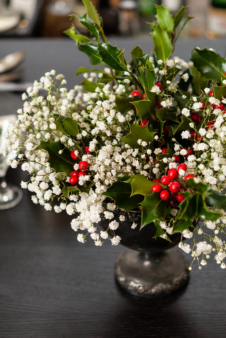 A silver vase with a holly and baby's breath bouquet on a dark wooden table.