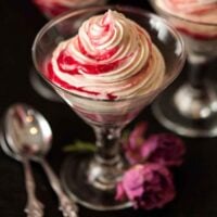 1 glass of white chocolate mousse with raspberry compote swirls on a black table with a few roses and spoons at their base.