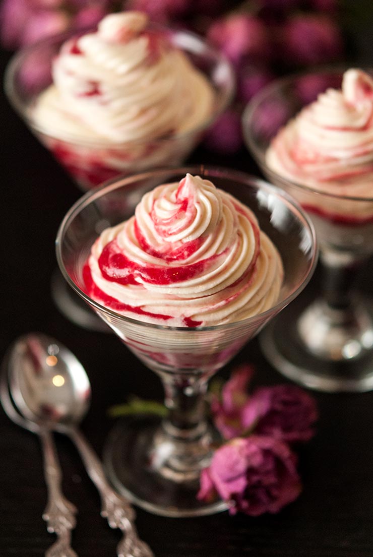 3 small glasses of mousse with raspberry compote on a table. 2 dry roses sit beside one glass, as well as 2 small spoons.