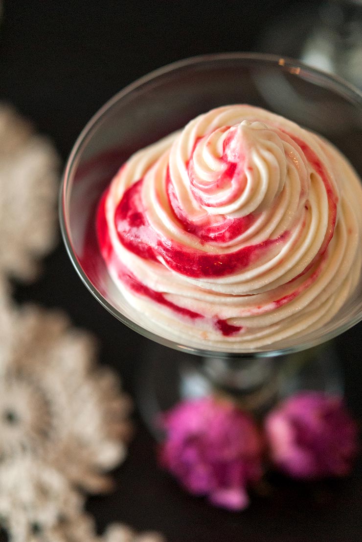 White chocolate mousse with raspberry swirls. 2 dry roses are seen below on a black table with lace.