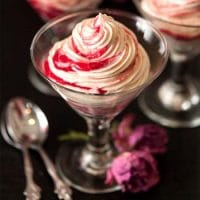 3 glasses of white chocolate mousse with raspberry compote swirls on a black table with a few roses and spoons at their base.