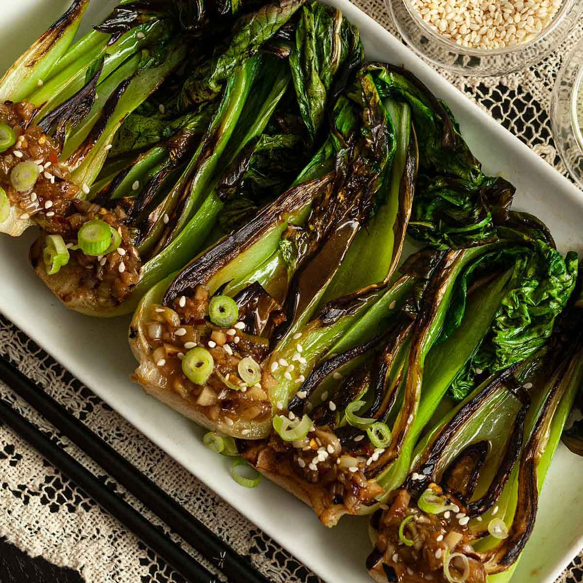 5 seared baby bok choy on a plate beside chop sticks, a bowl of sesame seeds and sauce on a lace table cloth.