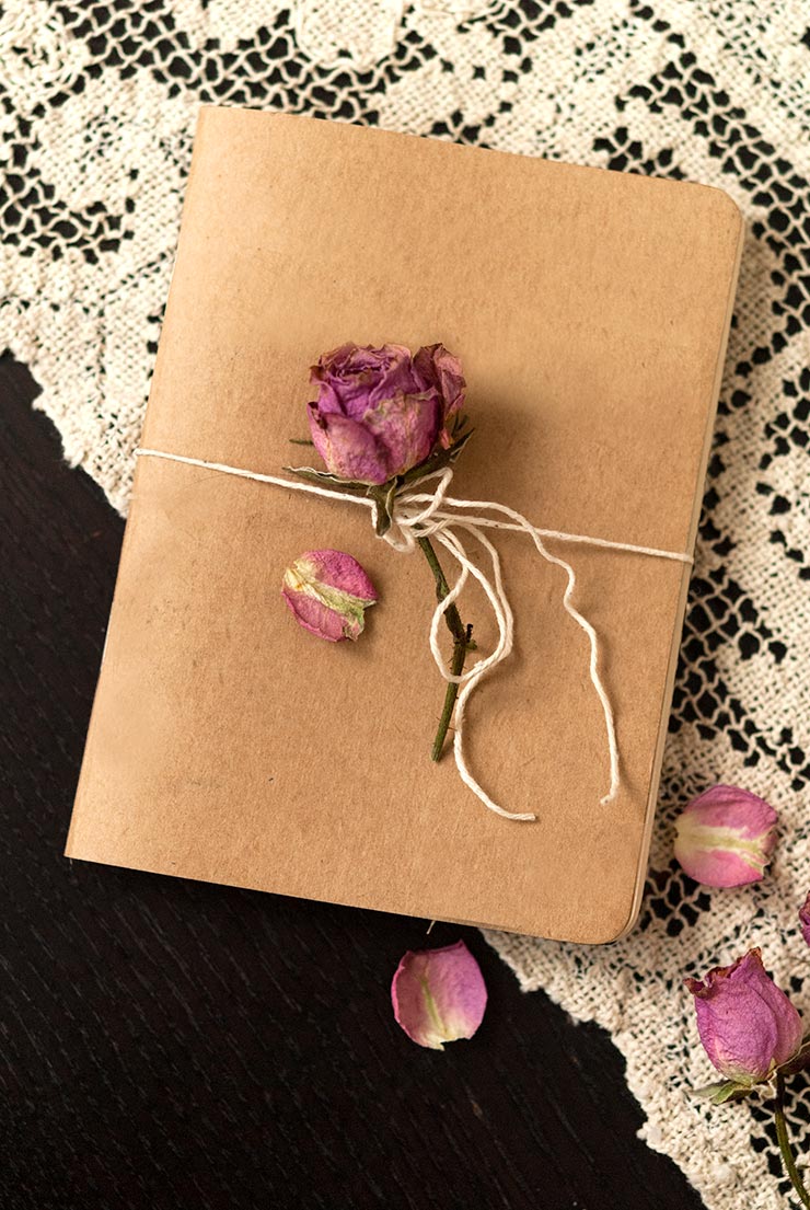 A small book tied with string and a rose, laying on a black table with a lace tablecloth, sprinkled with rose petals.