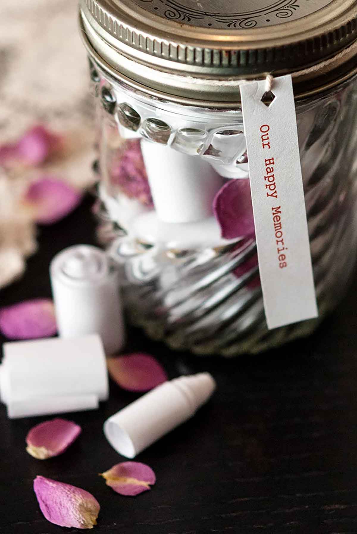 A jar with tiny scrolls inside and outside of it on a black table, sprinkled with rose petals.
