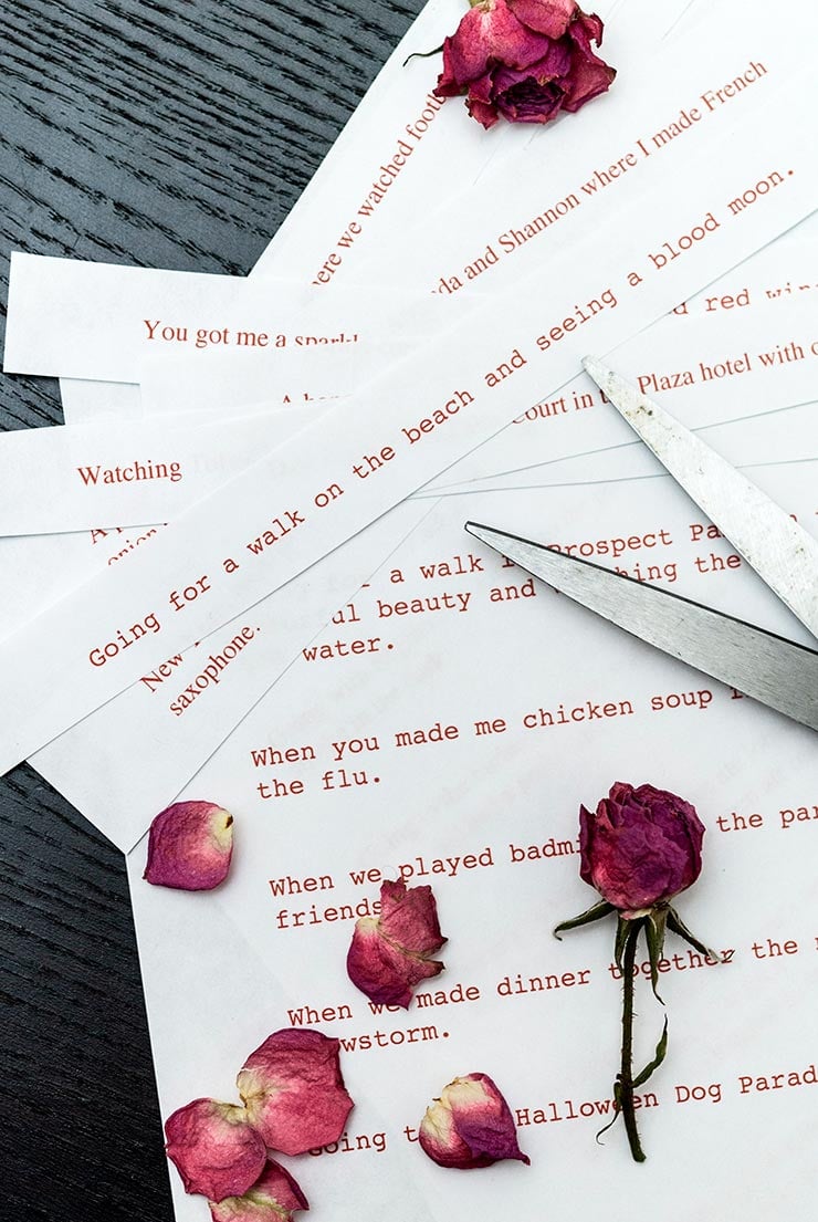 Strips of paper with memories written on them with a pair of scissors and a few rose petals on a black table.