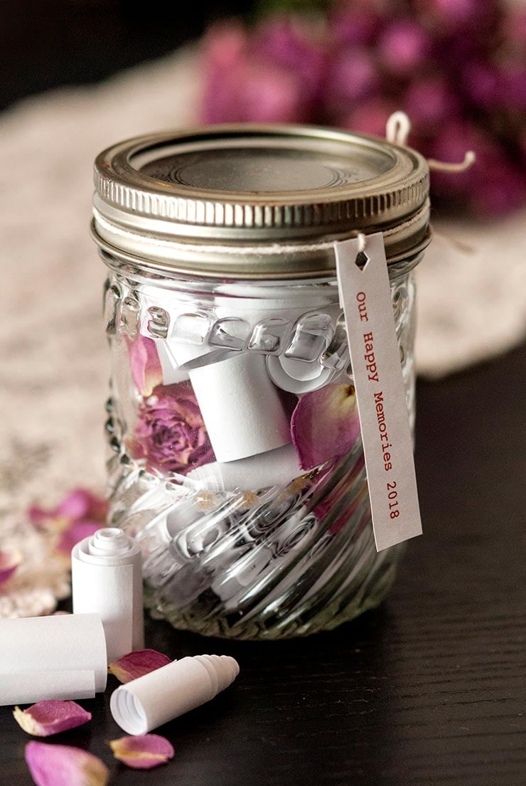 A jar filled with small scrolls and flower petals on a table, with a tag that reads "Our Happy Memories 2018."