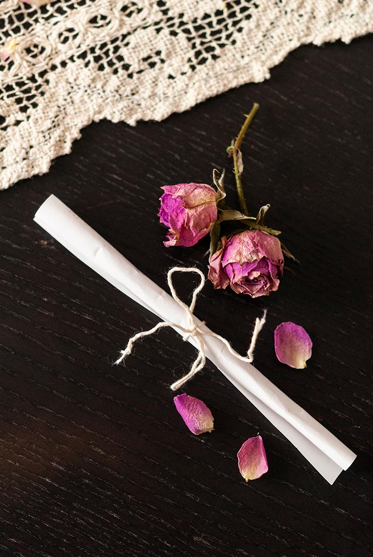 A small scroll of paper, tied with string, on a black table with sprinkled with rose petals beside a lace table cloth.