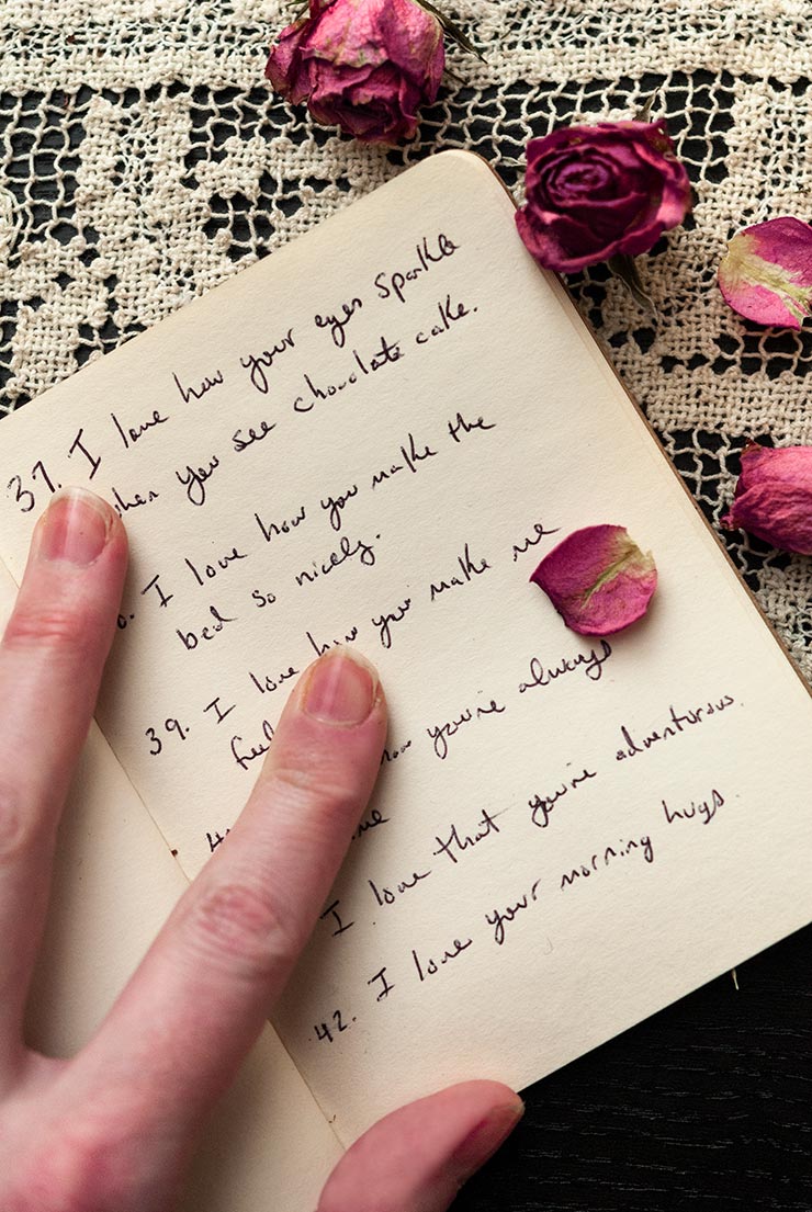 A hand holding open the pages of a book with writing inside on a lace table cloth, sprinkled with rose petals.