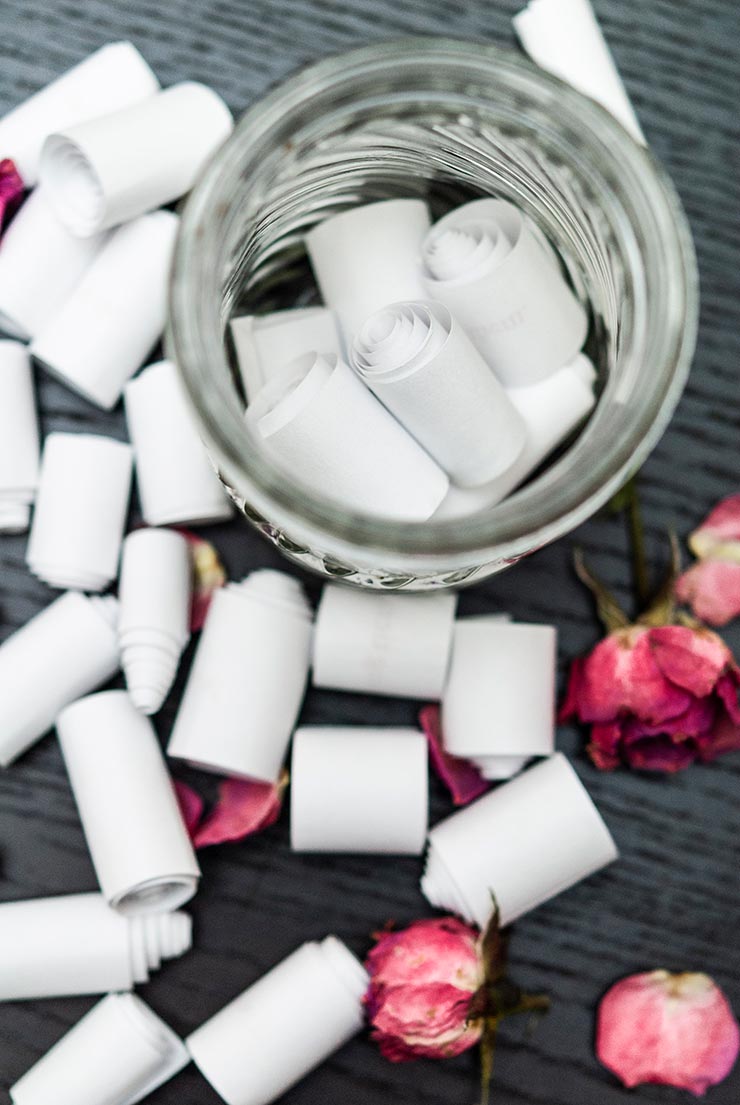 Little scrolls of paper outside of a jar, and a few inside, on a black table, sprinkled with rose petals.