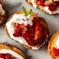 A balsamic strawberry crostini, garnished with a few small flowers, surrounded by others on a marble plate.