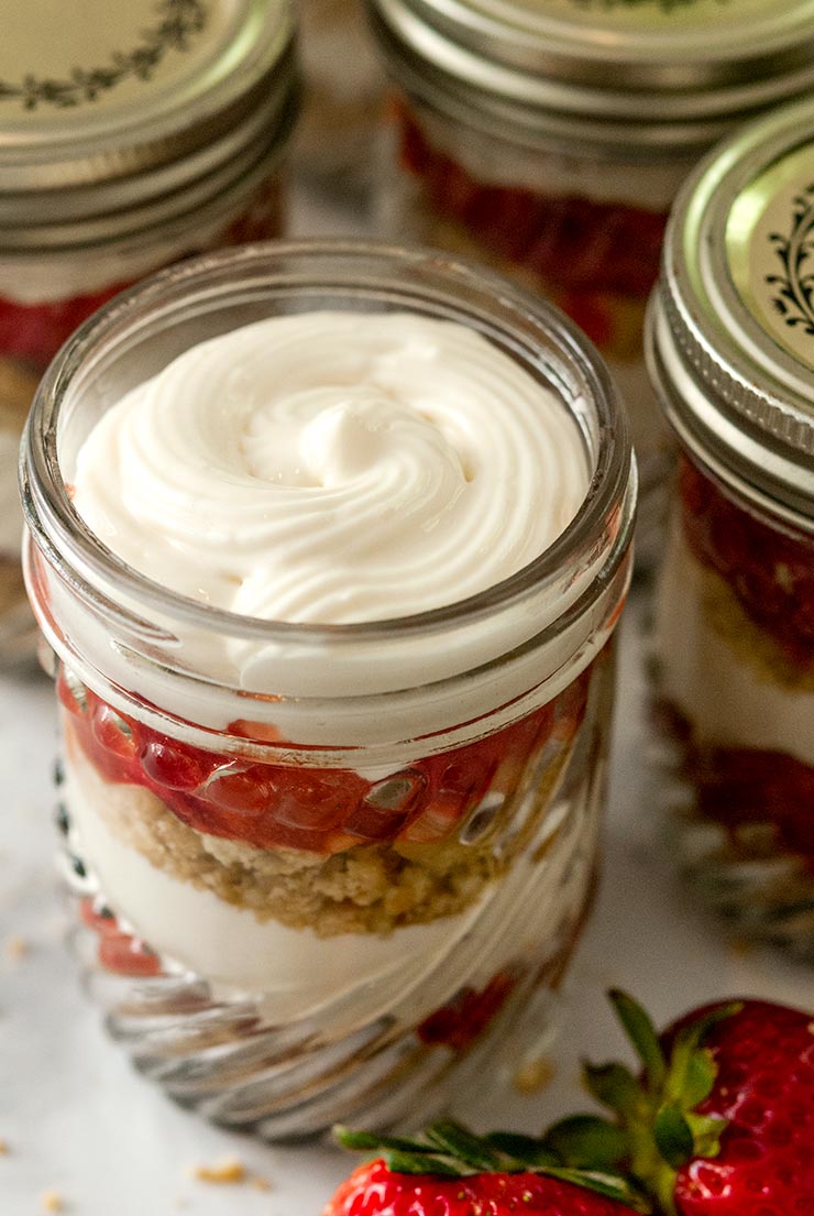 A strawberry rhubarb cup with vanilla cheesecake mouse on a marble table with a few strawberries and other jars beside it.