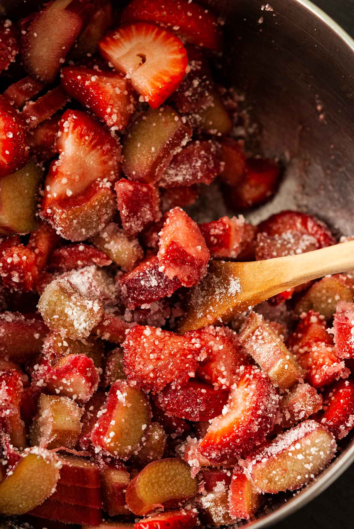 Raw strawberries and rhubarb in a mixing bowl with sugar and tapioca and a spoon.