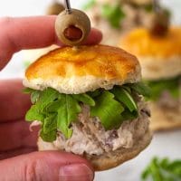 Fingers holding a chicken salad slider, up close, with other siders in the background.