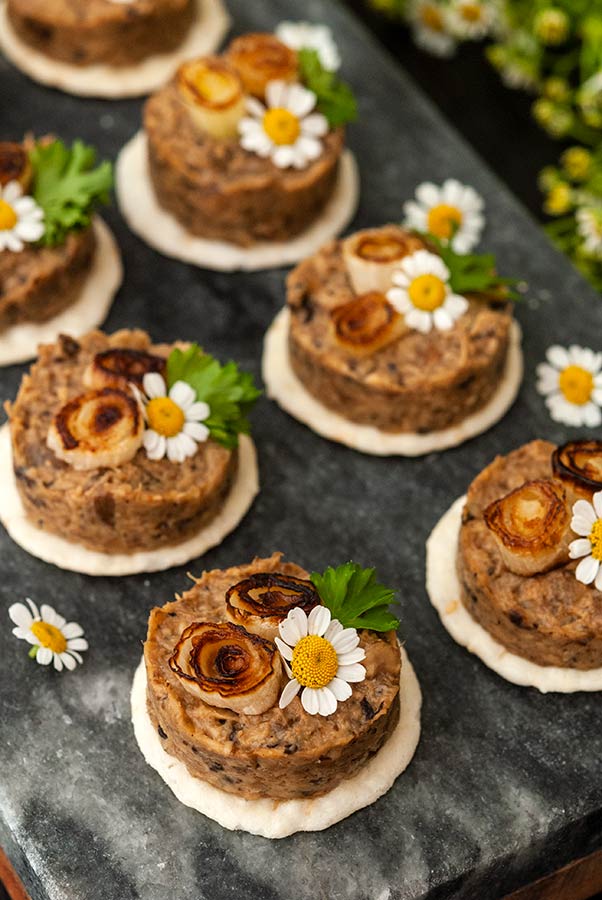 6 mushroom pâté canapés garnished with scallions and flowers on a marble slate.