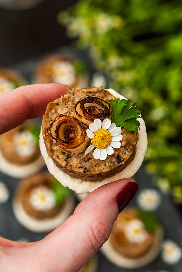 Fingers holding a mushroom pâté canapé above a plate of canapés and flowers.