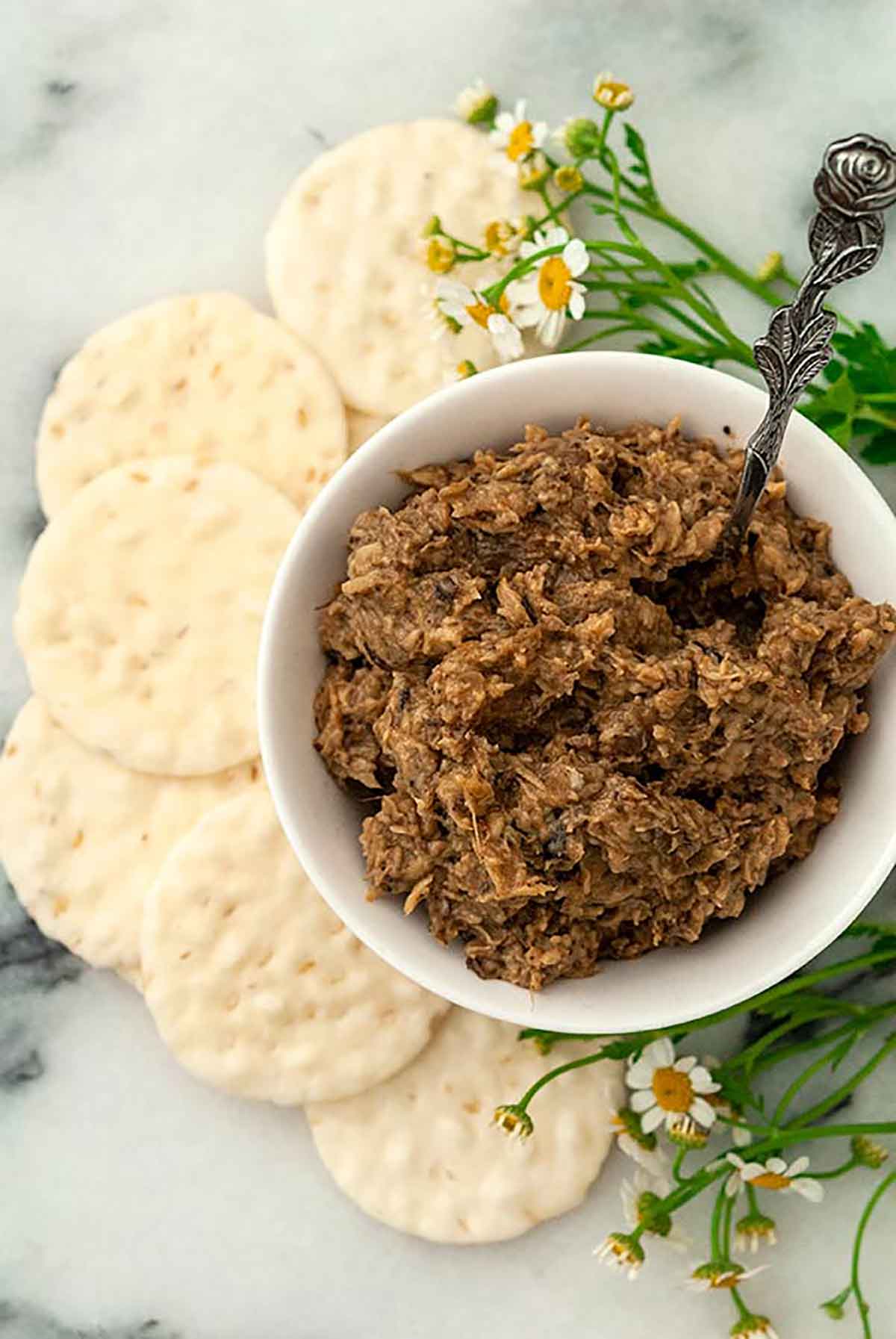 A bowl of vegan mushroom pâté surrounded by crackers and a few flowers.