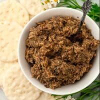 A bowl of mushroom pate, surrounded by rice crackers and green flower stems.