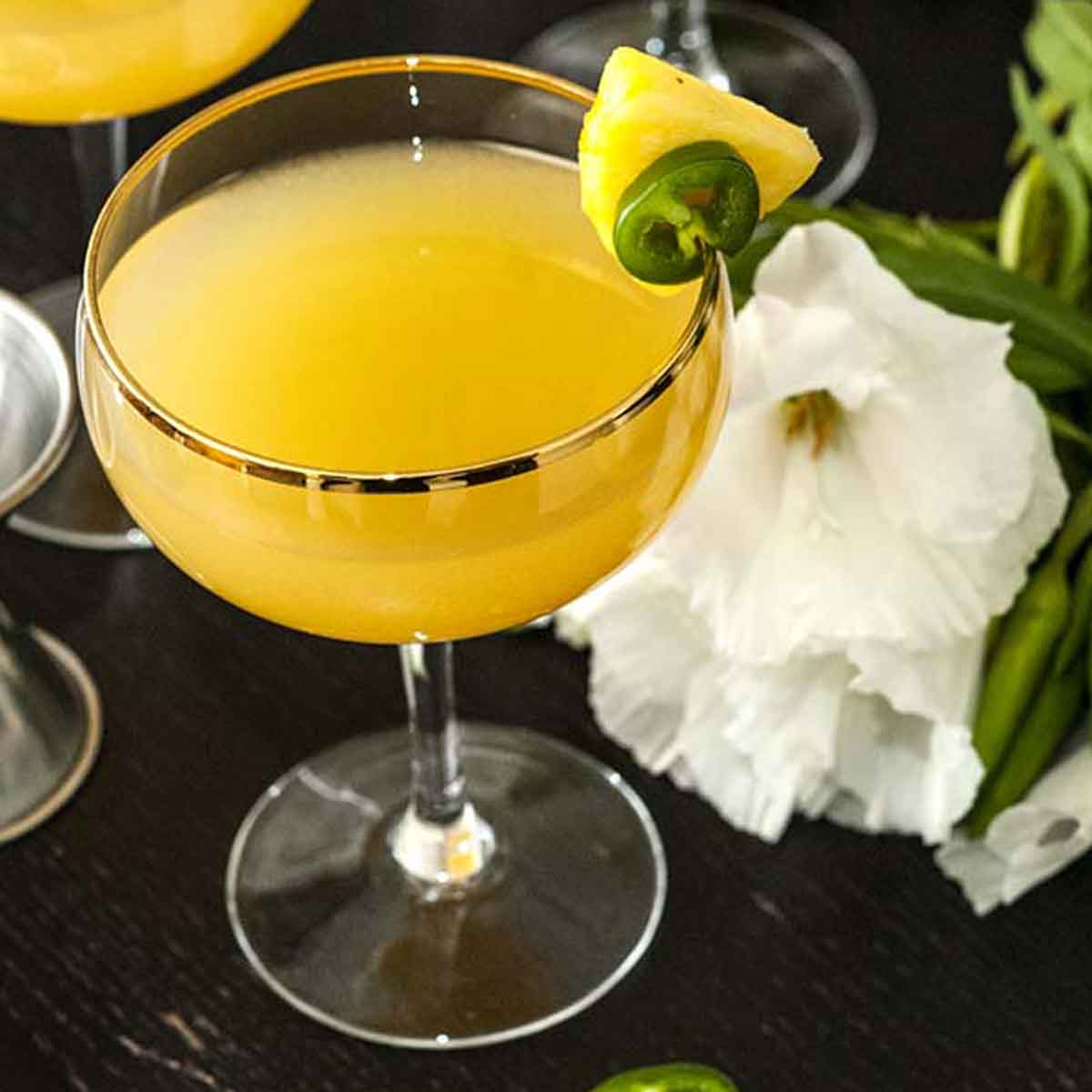 A pineapple cocktail on a table, garnished with pineapple and jalapeño, with tropical flowers in the background.