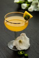 A pineapple cocktail on a table, garnished with pineapple and jalapeño, with tropical flowers in the background.