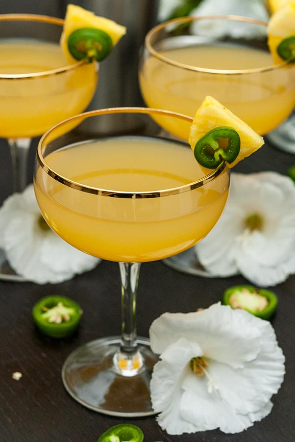 3 pineapple cocktails on a table, garnished with pineapple and jalapeños, surrounded by tropical flowers.