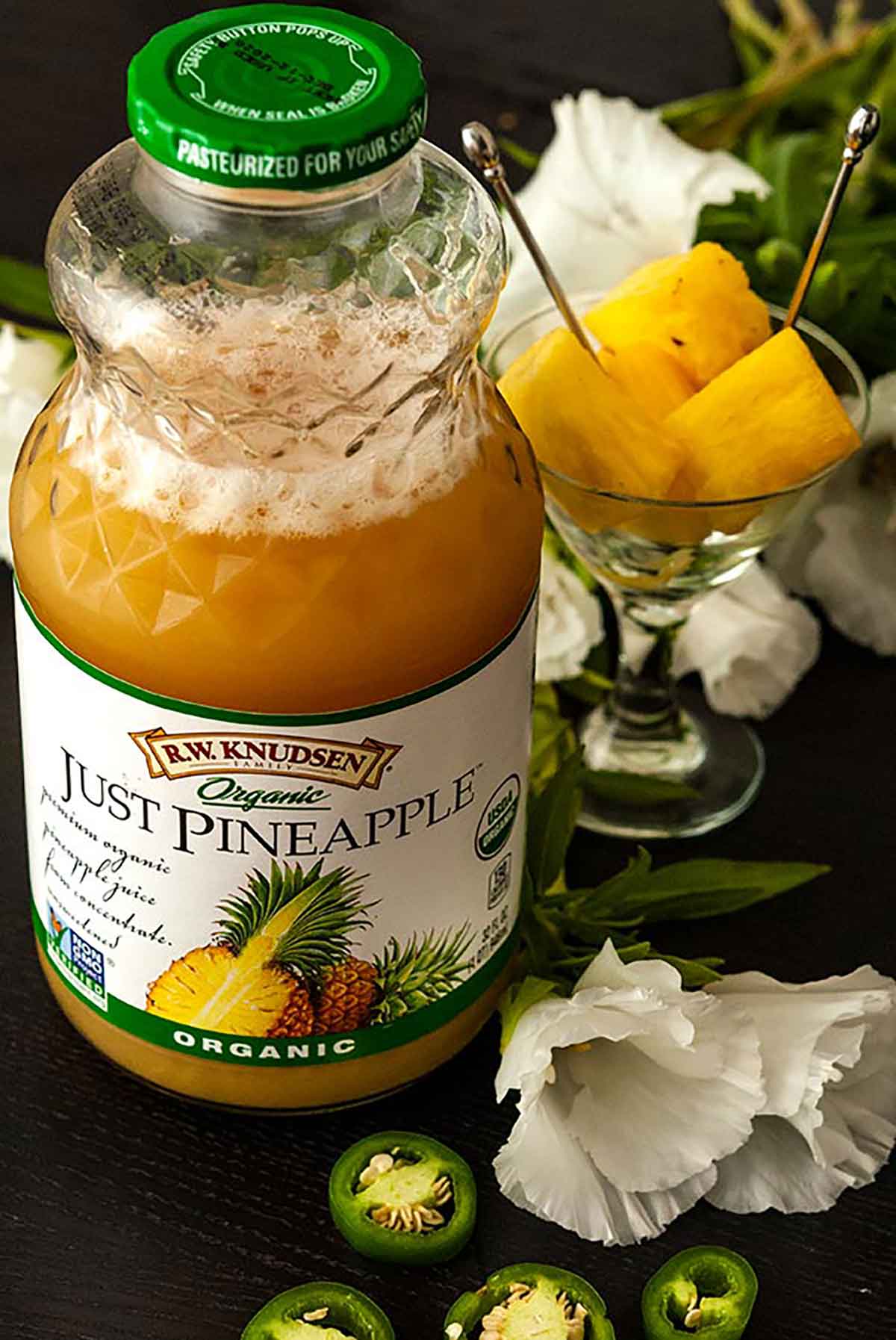 A bottle of pineapple juice on a table with flowers, jalapeños and a small glass of pineapple chunks.