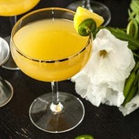 A pineapple cocktail on a table, garnished with pineapple and jalapeño, with tropical flowers in the background.