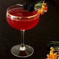 A cocktail on a wood table, garnished with flowers and blackberries.