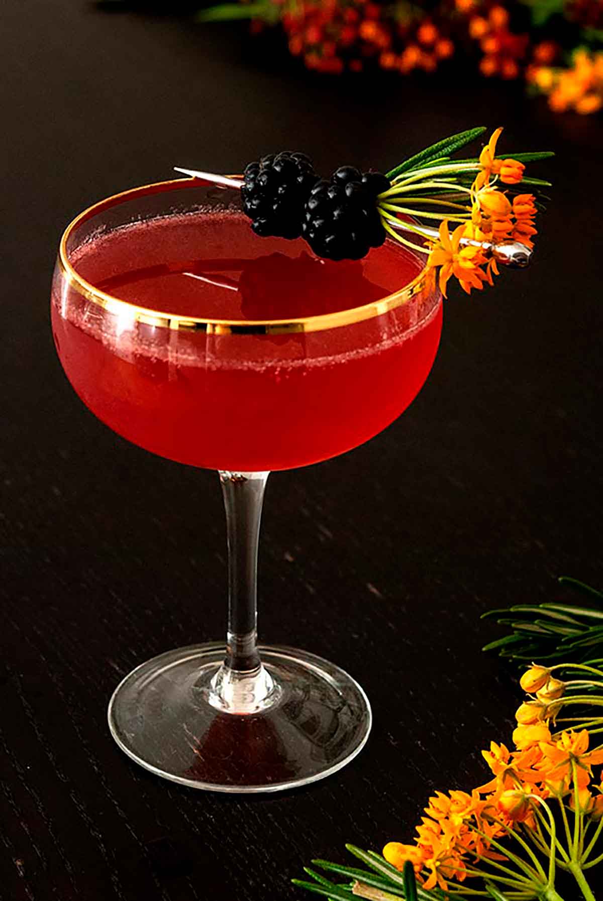 A cocktail on a wood table, garnished with flowers and blackberries.